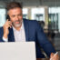 Businessman in a suit talking on the phone while working on a laptop in a modern office.